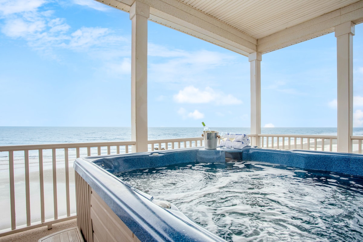 A spacious hot tub is showcased on an upper deck, with views of the ocean visible beyond. The tub is filled with bubbling water and surrounded by a protective railing, while a cooler with refreshments and towels can be seen nearby.