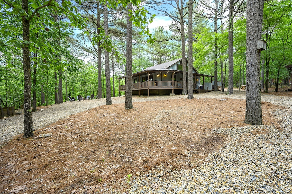 The cabin is surrounded by tall pine trees and sits on a naturally landscaped area covered with pine needles. A wrap-around porch offers multiple entry points, while a gravel path leads to a charming seating arrangement near the fire pit.