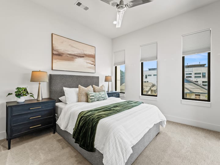 Queen bedroom with dark upholstered headboard, desert landscape artwork & sage green accent throw — a warm, stylish retreat. Ceiling fan & soft lamp lighting for total comfort.