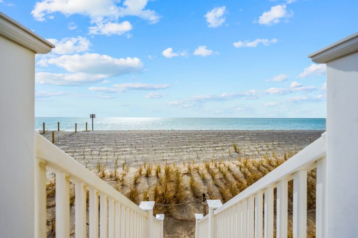 A clear view of the ocean is seen from the top of a set of white stairs, leading down to a sandy beach area. The sky is bright with scattered clouds, reflecting a tranquil atmosphere. Tall grasses frame the sandy path towards the water.