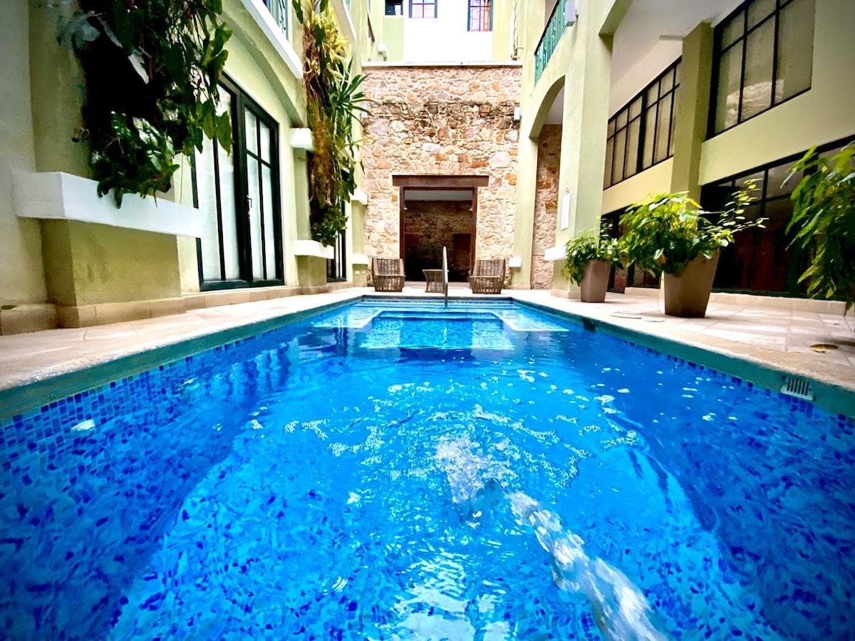 A serene communal pool is featured, surrounded by tall plants and smooth stone walls. The water reflects an inviting blue hue, while two lounge chairs are positioned nearby, offering a space for relaxation. Bright natural light illuminates the area, enhancing the tranquil setting.