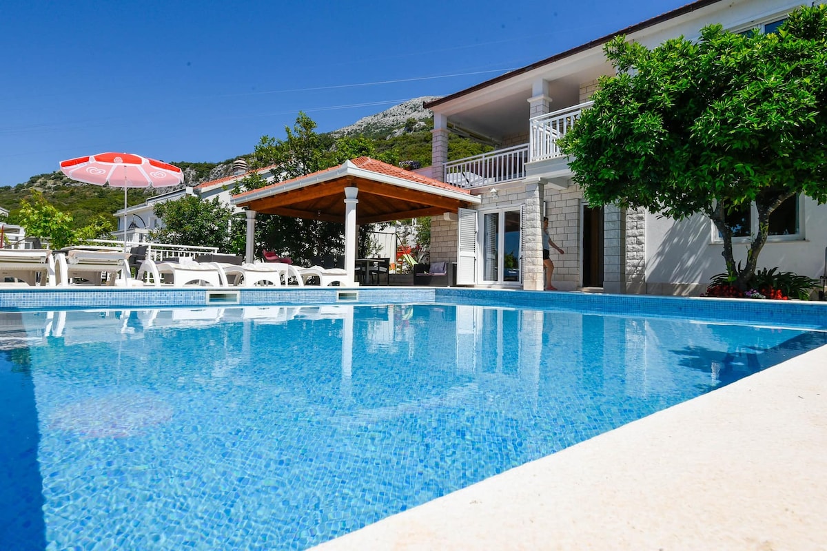 A clear swimming pool reflects the blue sky, surrounded by a spacious terrace with sun loungers and an umbrella. A covered patio area is visible, along with the modern facade of the villa in the background, framed by greenery.