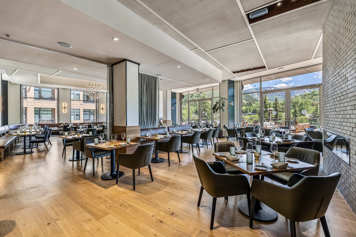 The dining area features a modern design with wooden flooring and large windows offering views of the outdoors. Tables are set for dining, with comfortable chairs arranged symmetrically. Soft lighting from overhead fixtures enhances the inviting atmosphere.