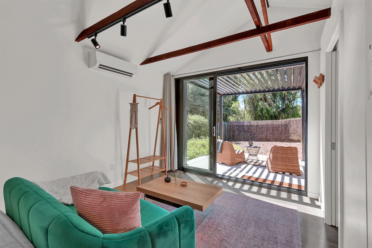 A modern living area features a green loveseat and a light wooden coffee table. Large glass doors open to an outdoor patio with seating surrounded by lush greenery, allowing for natural light to fill the space.