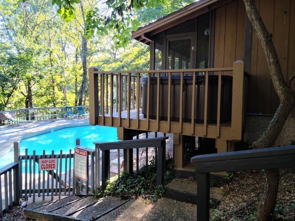 A wooden deck leads to a charming cottage, with a screened-in porch visible through the glass door. A semi-private pool is surrounded by trees, and a 'Keep Area Closed' sign indicates the space for guest safety.
