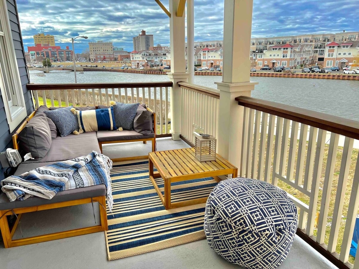 A covered porch area features a comfortable seating arrangement with plush cushions and throws. A wooden coffee table sits between two seating sections on a decorative rug. A compact pouf adds a relaxing touch, with views of the water and surrounding buildings in the background.