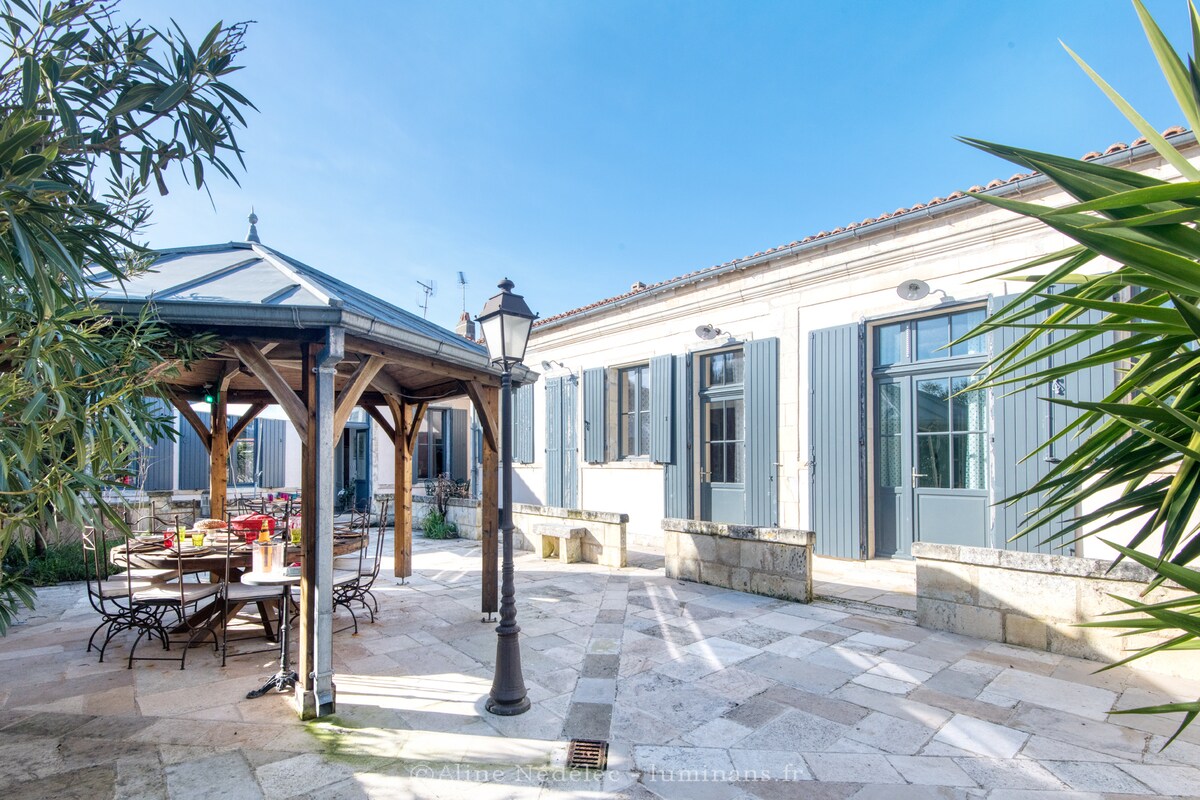 A spacious outdoor courtyard is featured, with a wooden gazebo providing shade over a dining table set for guests. Surrounding the area are well-maintained plants and large windows of the house, allowing natural light to fill the space.