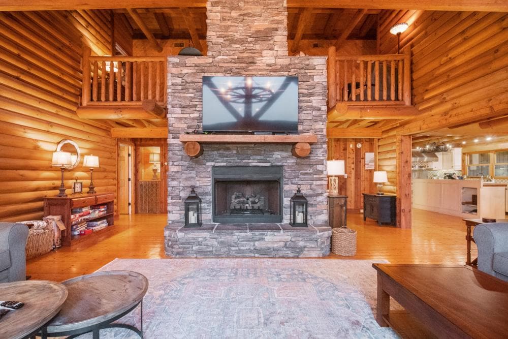 A spacious living area features a large stone fireplace as a focal point, with a flat-screen TV mounted above. Cozy seating arrangements are positioned around a patterned area rug, while warm wood beams and walls create a rustic ambiance. Soft lighting illuminates the inviting atmosphere.