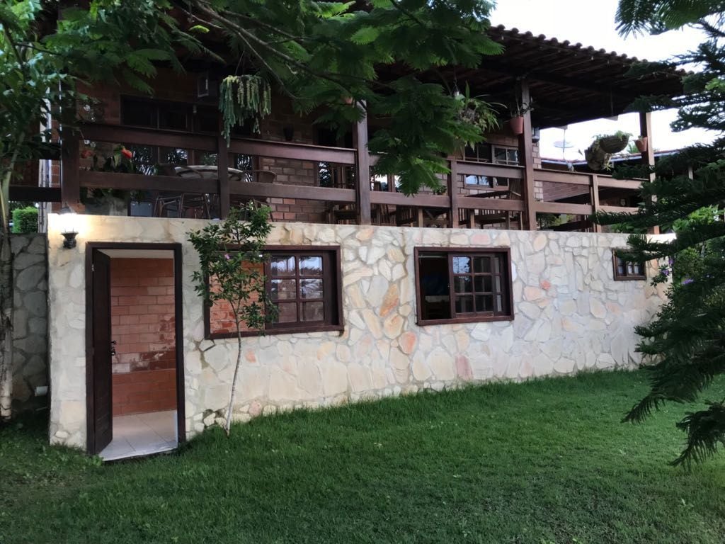 A multi-level house is viewed from the landscaped yard, framed by lush greenery. The exterior combines natural stone and wooden elements, showcasing spacious balconies with wooden railings. Windows are set into the stone facade, providing a harmonious connection with the surrounding environment.