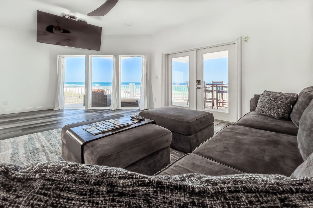 A spacious living room features a dark sectional sofa complemented by a textured area rug. Large glass doors provide a panoramic view of the ocean, allowing natural light to fill the space while connecting to the private beachfront deck.