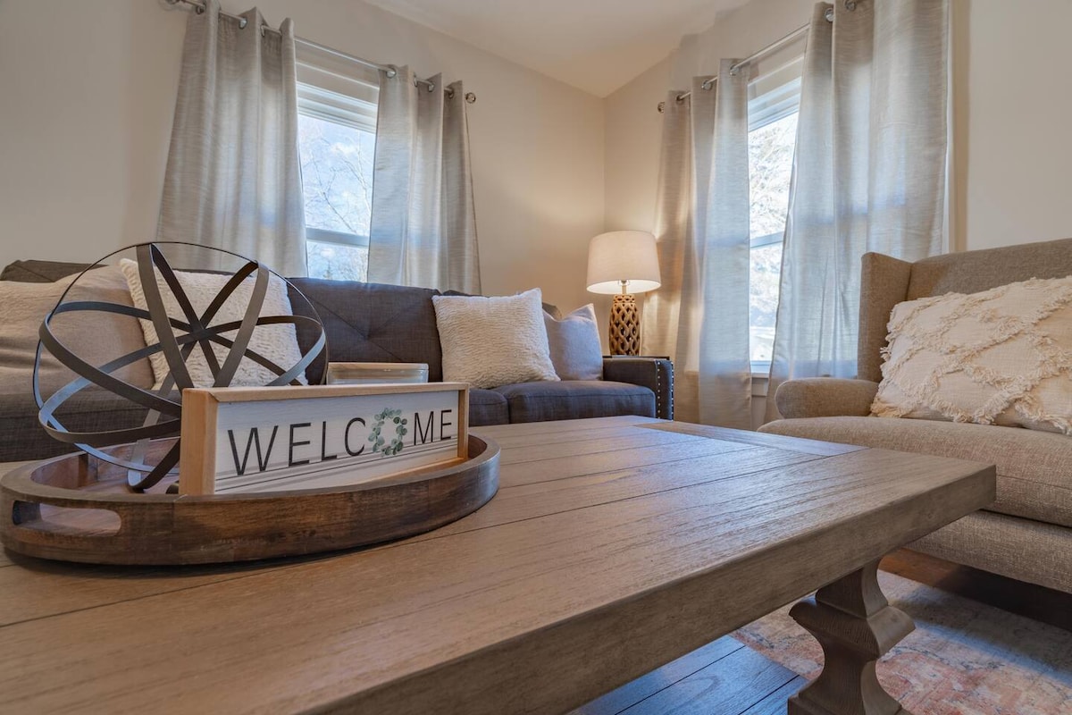 A cozy living area is centered around a wooden coffee table with a welcome sign. Two sofas, one in gray and another in a soft beige, are positioned around the table. Natural light filters through curtains at the windows, creating a warm atmosphere.