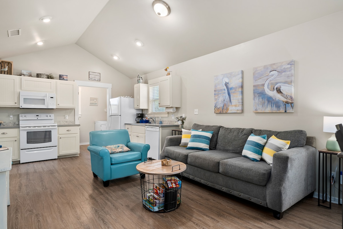 The inviting living area features a comfortable gray sofa with colorful accent pillows opposite a light wood coffee table. A crown of coastal-themed art adorns the walls, and a turquoise armchair adds a pop of color. A fully equipped kitchen is visible in the background.