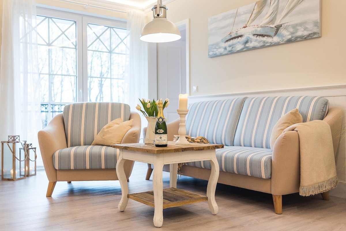 A welcoming living area features soft striped sofas positioned around a wooden coffee table. A vase of yellow tulips adds a touch of color. Large windows offer plenty of natural light, complemented by a pendant light overhead.