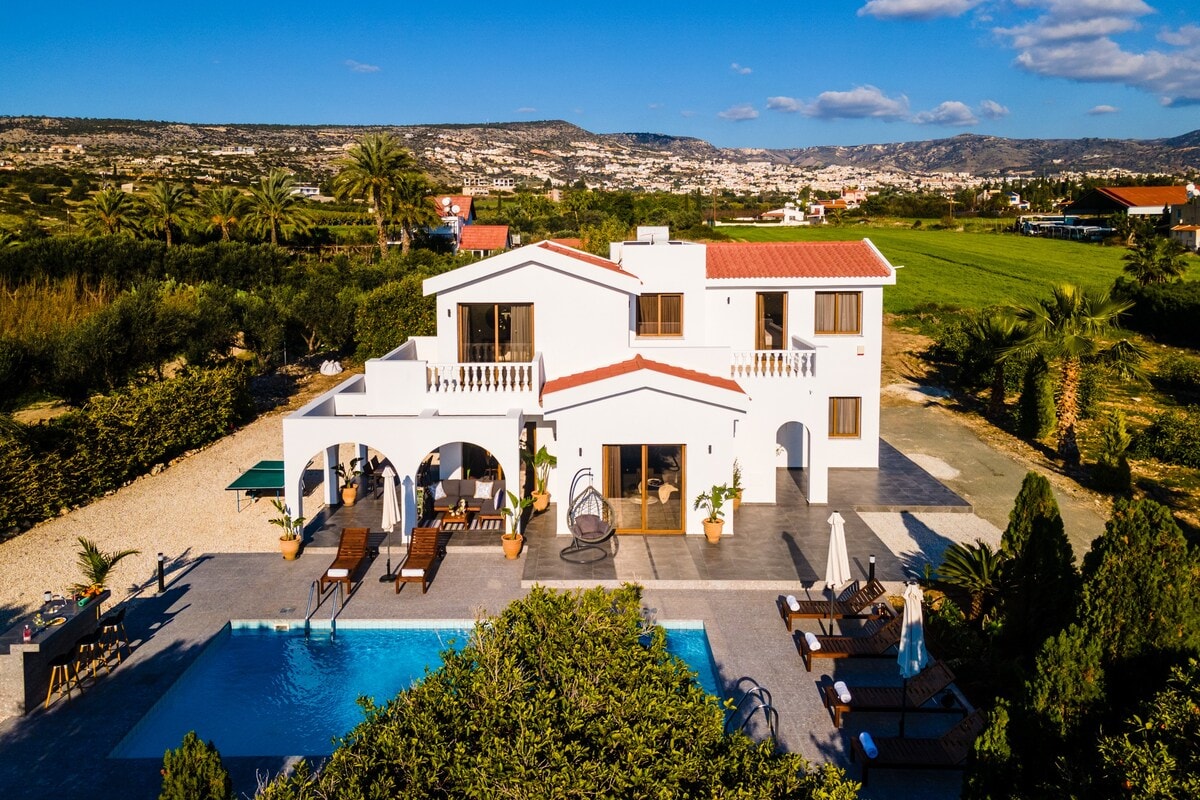 The villa features a spacious outdoor area with a large pool surrounded by sunbeds and tropical landscaping. A covered patio is visible, offering seating for relaxation. The two-story structure displays a contemporary design with large windows and a rooftop terrace, overlooking scenic countryside and distant hills.