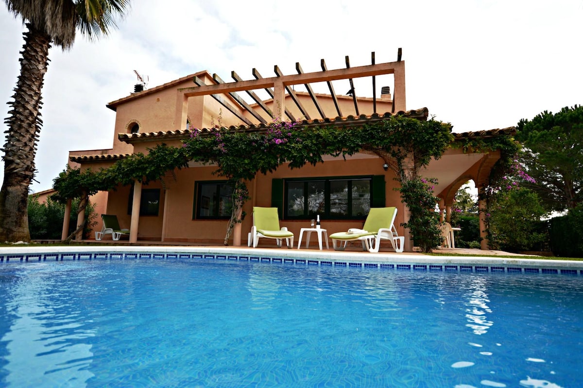 An outdoor view of a vacation home featuring a swimming pool in the foreground. The house is adorned with greenery and a trellis, providing shade. Two light-colored lounge chairs are positioned by the pool, creating a relaxing atmosphere in the garden.