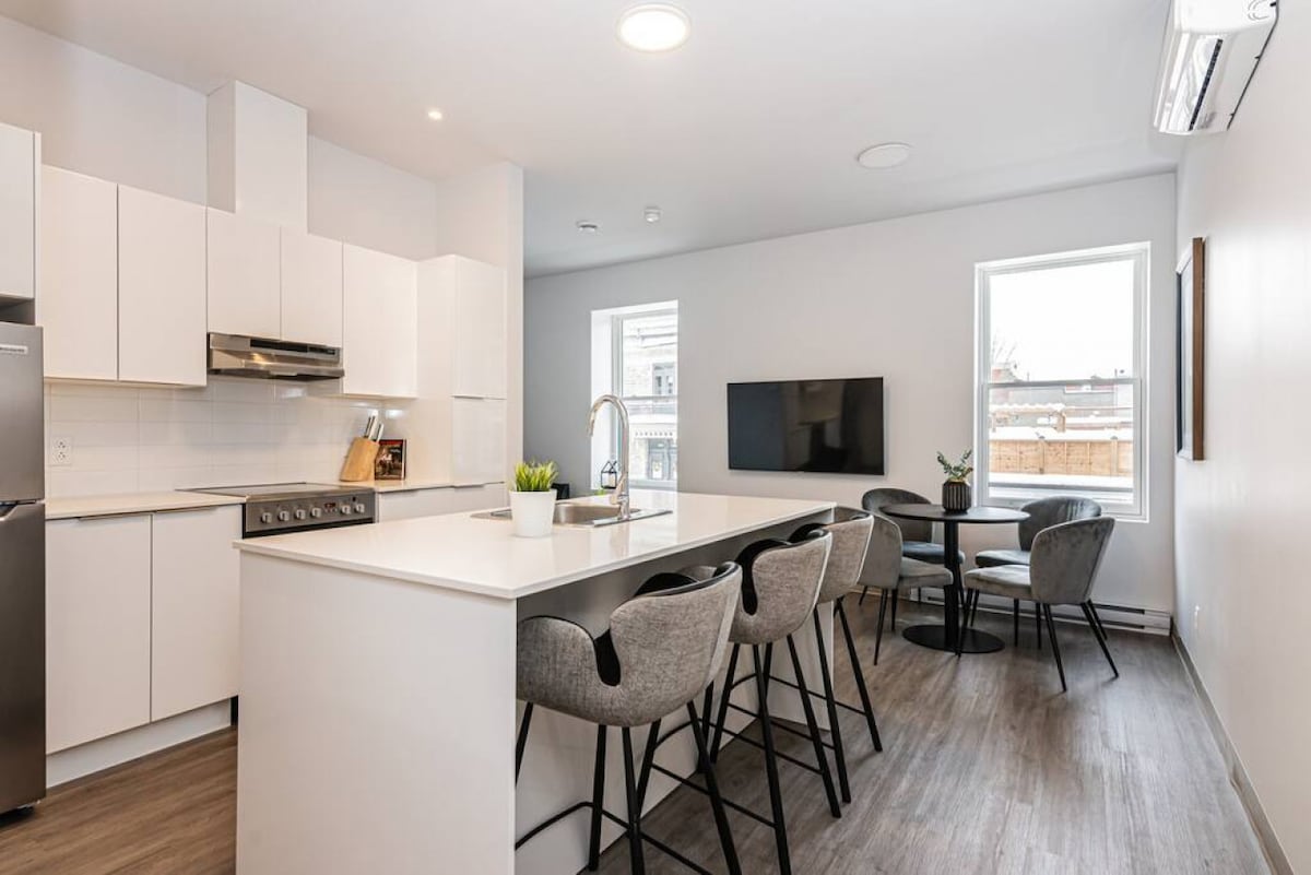 A bright kitchen and dining area features modern white cabinetry and high-end appliances. A spacious island with seating is complemented by a round dining table surrounded by four chairs. Two windows provide natural light, and a wall-mounted TV enhances the entertainment options.