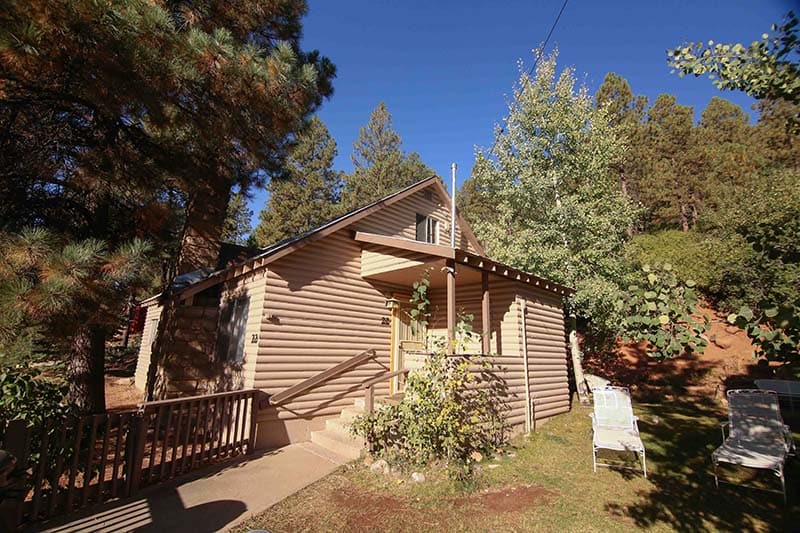 The exterior of a rustic cabin is depicted, showcasing a well-maintained façade surrounded by trees. A set of stairs leads to the entrance, with inviting greenery and shrubs along the pathway. Two lounge chairs are positioned nearby, creating a comfortable outdoor seating area.