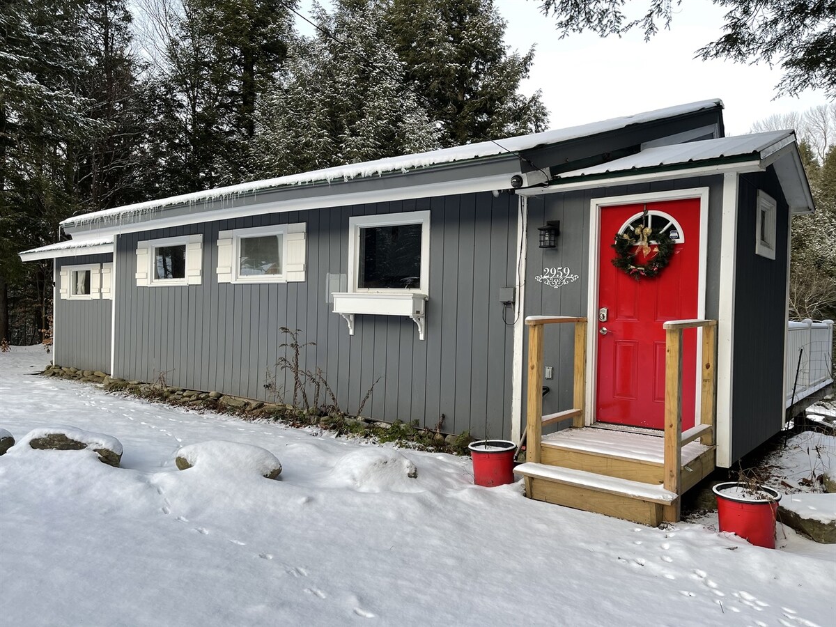 The exterior of the chalet is depicted, featuring a grey facade with multiple windows. A bright red door is accented by a seasonal wreath. Surrounding areas are covered in snow, and potted plants are positioned near the entrance, contributing to a welcoming appearance.