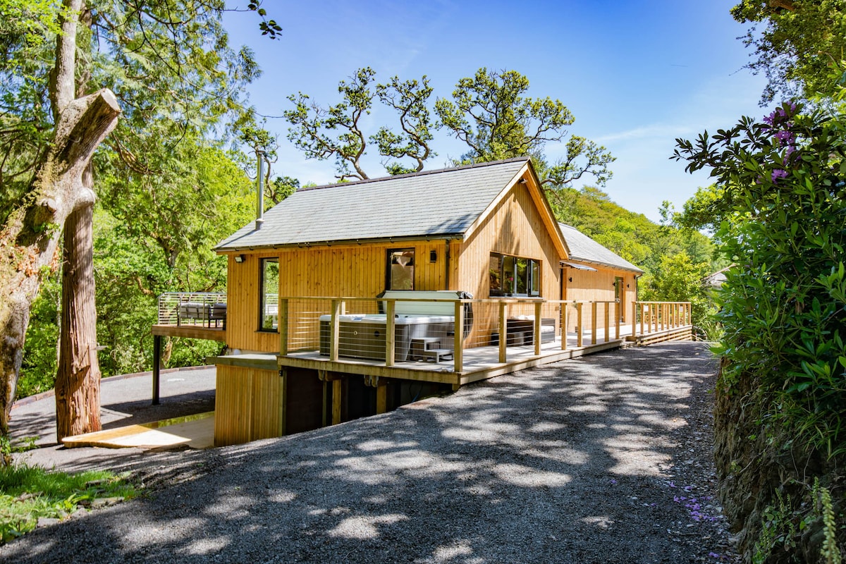 A modern lodge constructed from natural wood is depicted, nestled amidst lush greenery. A spacious deck extends from the lodge, providing an outdoor space for relaxation. The sunlit facade showcases large windows that invite light into the interior, surrounded by trees casting dappled shadows.