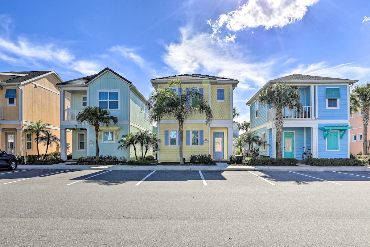 A two-story vacation home is displayed among colorful neighboring houses. Palm trees line the property, providing greenery. The building's exterior features bright yellow siding with large windows, allowing natural light to fill the interior. A parking area is visible in front of the homes.