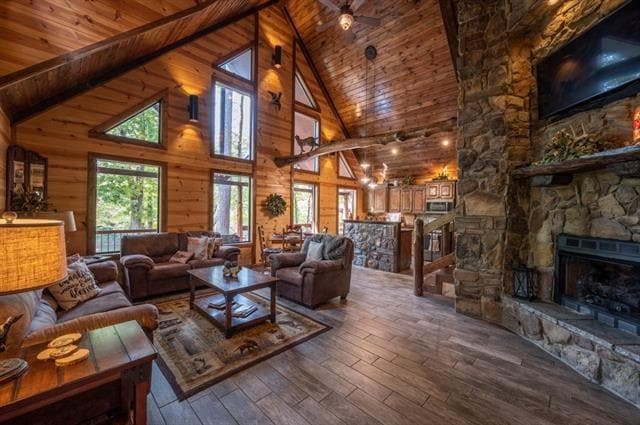 A spacious living area features vaulted wood ceilings and large windows, allowing natural light to fill the room. Comfortable seating is arranged around a coffee table, with a stone fireplace providing a cozy focal point. The open layout leads towards the kitchen area in the background.