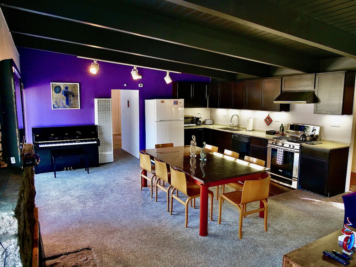 An open-concept kitchen and dining area features a large table with several chairs, surrounded by modern cabinets and appliances. A piano is positioned to the left alongside a wall adorned with a piece of art. Recessed lighting illuminates the space, enhancing the vibrant purple walls.