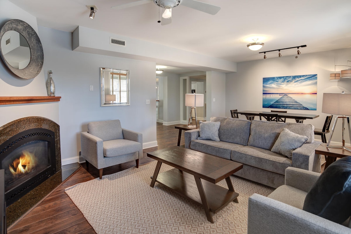 A light-filled living area is featured, showcasing a comfortable gray sofa and two armchairs arranged around a wooden coffee table. A stone fireplace provides warmth, while a large piece of wall art depicting a serene beach scene adds a calming touch. A dining area is visible in the background.