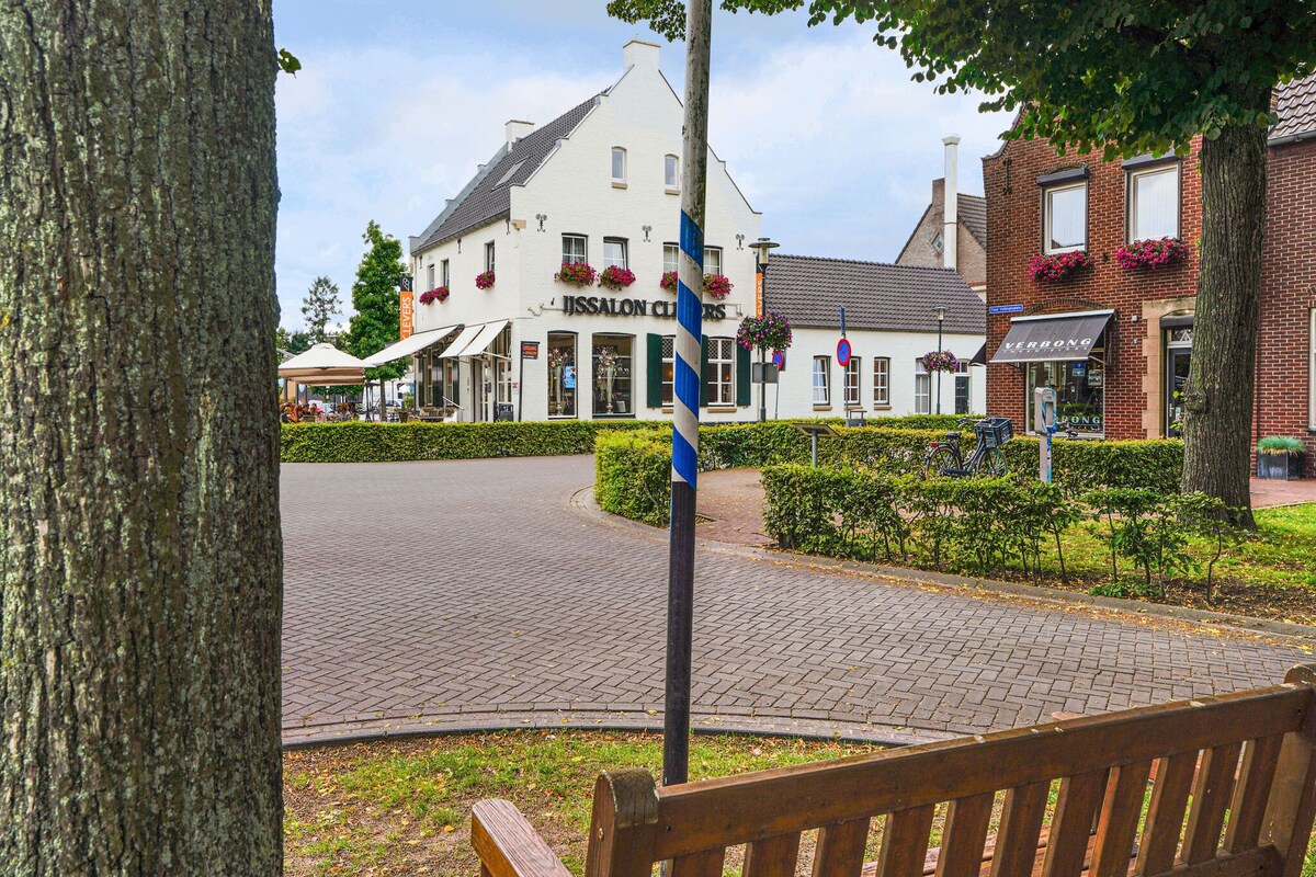 A serene street view is captured, showcasing a charming ice cream parlor and surrounding buildings. Benches and well-maintained green hedges create an inviting outdoor space. Trees provide shade, enhancing the tranquil atmosphere. Cobblestone paths can be seen leading further into the neighborhood.