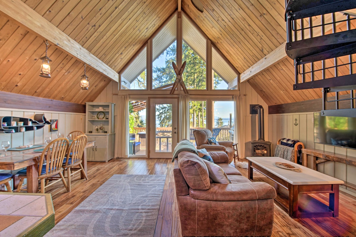 A cozy living area features high wooden ceilings and large windows that allow natural light to fill the space. Comfortable seating is arranged around a central coffee table. A dining table is visible in the background, set against a wall of light wood paneling.