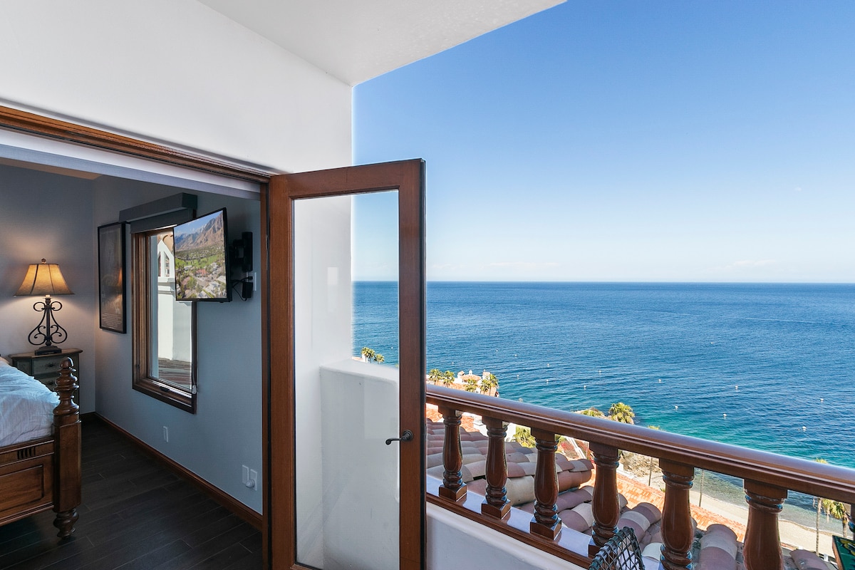 A private balcony offers sweeping views of the ocean, with bright blue waters stretching to the horizon. An open door reveals a glimpse of the bedroom, complemented by light streaming in from the window. The space is relaxed and inviting.