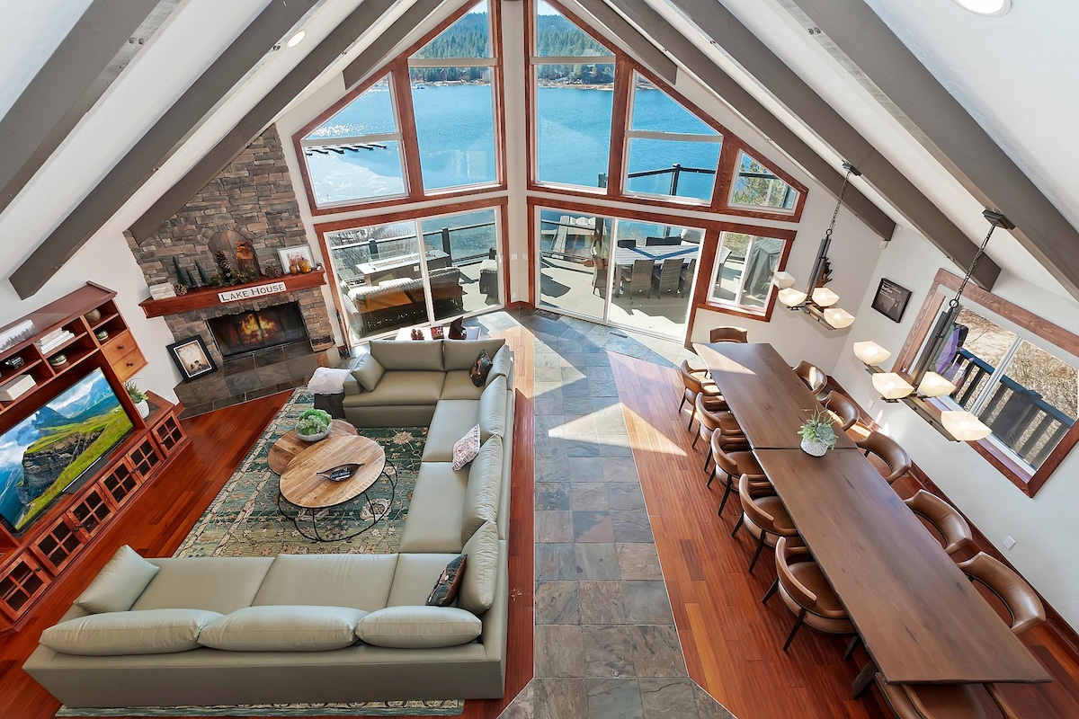 The spacious living area features high ceilings with exposed beams and floor-to-ceiling windows, offering panoramic views of the lake. A large sectional sofa surrounds a wooden coffee table, while a dining area with a long table accommodates numerous guests. A stone fireplace adds warmth to the room.