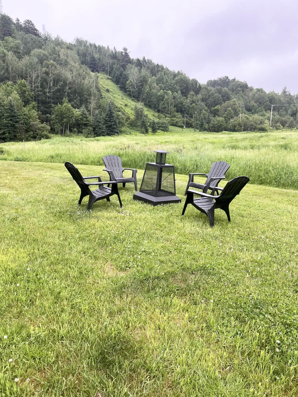 Four black outdoor chairs are arranged around a central fire pit on a grassy area. Lush green hills and trees surround the space, providing a natural backdrop. The scene appears peaceful, ideal for gatherings or relaxation.