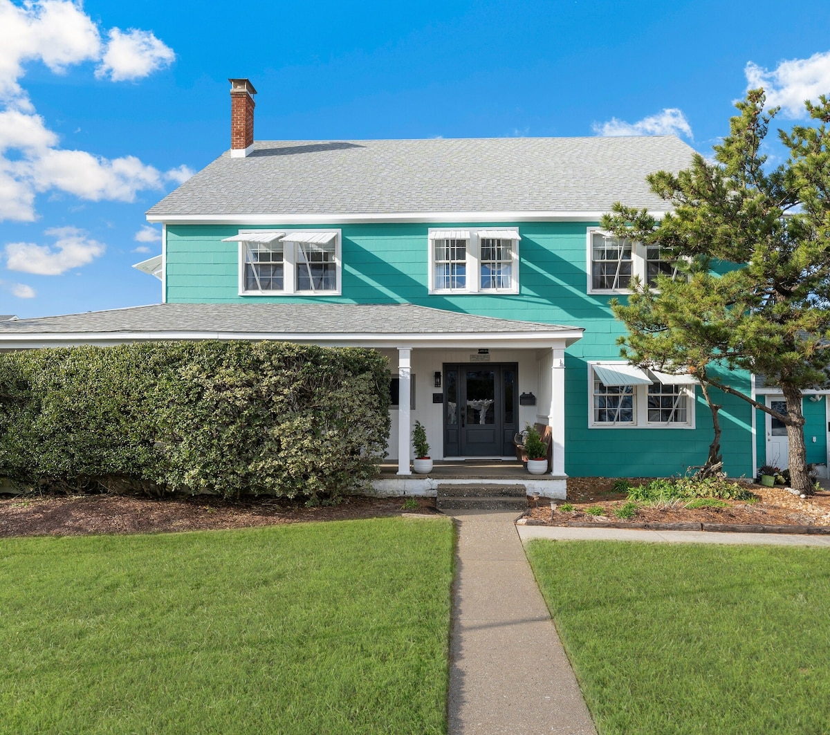 A two-story building with teal siding is showcased, featuring white trim around the windows and a welcoming front door. A manicured lawn lines the pathway leading to the entrance, framed by greenery and bushes, while a large tree adds to the charm of the exterior.