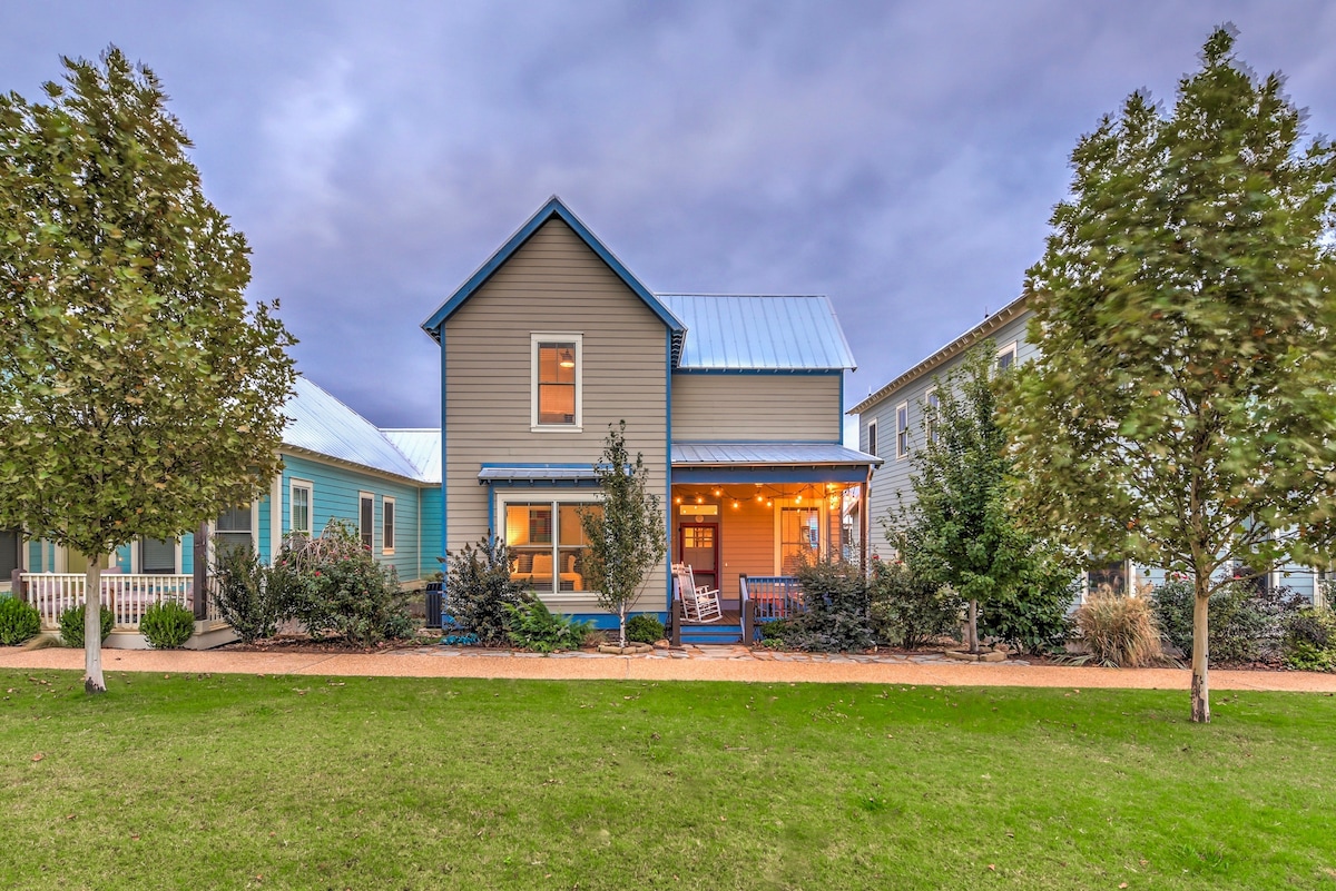 The exterior of Poindexter Cottage is captured at dusk, showcasing the two-story structure with a light blue roof and welcoming porch. Lush landscaping and shrubs frame the front, while a grassy area invites relaxation. Soft lighting from the porch creates an inviting ambiance.