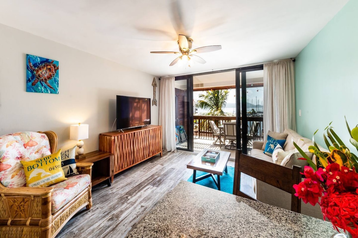 A bright living room features a ceiling fan and comfortable seating arrangements. Large glass doors provide access to a balcony that overlooks the marina. The space includes a television and a decorative side table, complemented by tasteful wall art and tropical floral arrangements.