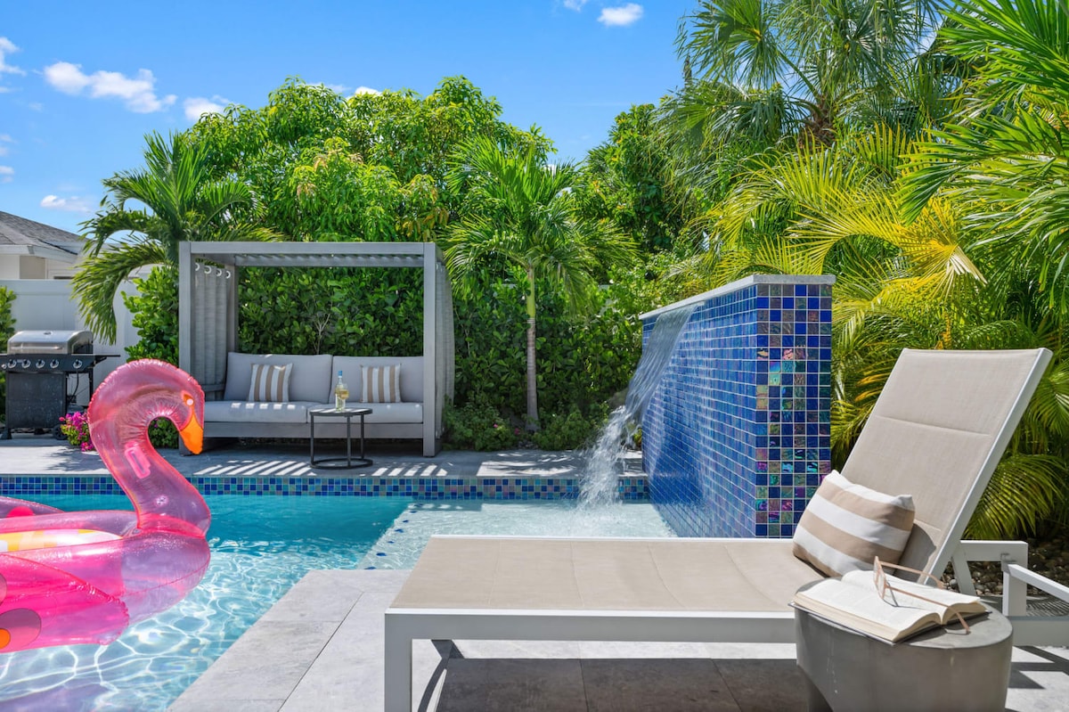 A serene backyard oasis is depicted, featuring a heated pool with a vibrant pink flamingo float. A modern lounge chair is positioned nearby, complemented by a side table and a book. A stylish gazebo offers additional seating, surrounded by lush tropical foliage and a waterfall fountain.