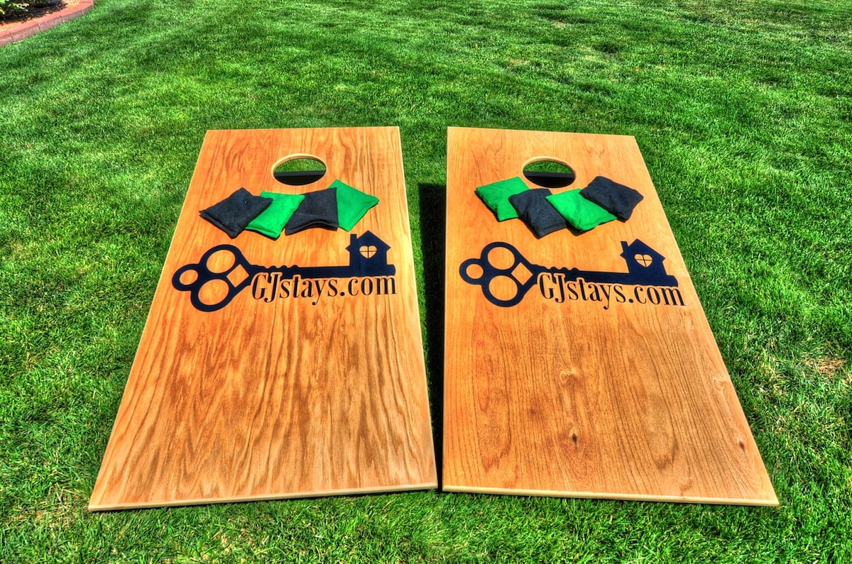 Two cornhole boards are set up on a green lawn, featuring a smooth wooden surface and an outlined hole in the center. Each board is adorned with the logo 'CjStays.com' and accompanied by four bags in green and black, neatly placed on each board.