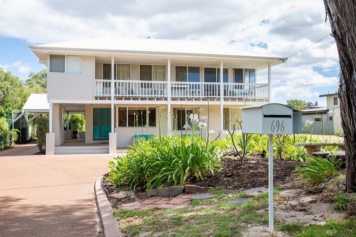 A two-storey coastal retreat with a light-colored exterior is surrounded by lush greenery. The front porch is lined with sliding glass doors, allowing for natural light. A mailbox is visible on the right, marked with the number 696, contributing to the welcoming entryway.