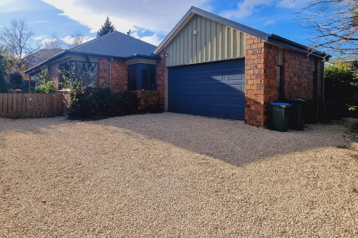 A single-story brick home with a combination of stone and metal siding is showcased. A spacious gravel driveway leads to a double garage with a blue door. Surrounding greenery adds a natural element, enhancing the curb appeal of the property.