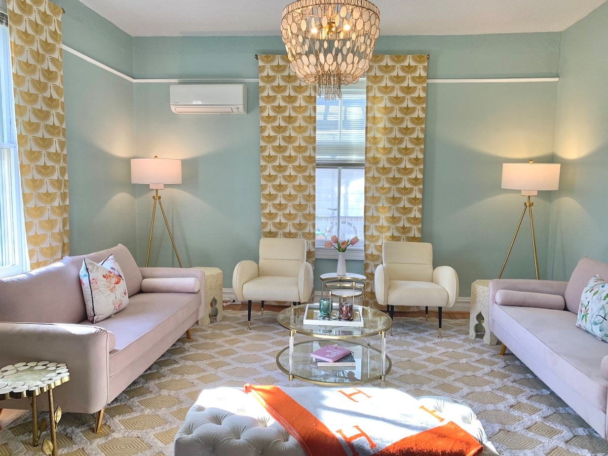 A bright living room features two pastel-toned sofas flanking a glass coffee table. Elegant armchairs are positioned nearby, illuminated by soft lighting from modern floor lamps. Textured walls and yellow patterned curtains enhance the inviting atmosphere, while a geometric area rug adds warmth underfoot.