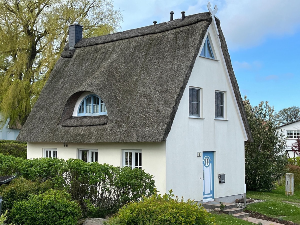 A charming thatched-roof holiday home is presented, featuring a bright white exterior with distinctive rounded windows. A welcoming blue front door and manicured greenery surround the entrance, highlighting a serene, nature-infused environment ideal for relaxation.