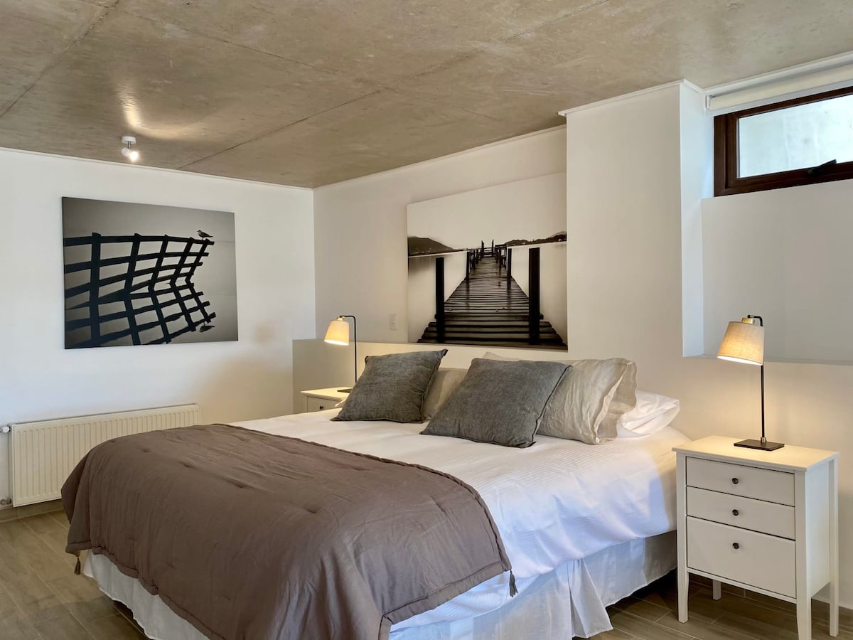 A spacious sleeping area features a large bed with a neutral-toned blanket and decorative pillows. Two bedside tables with lamps flank the bed. Large artwork adorns the walls, and natural light filters through a window, enhancing the room's relaxed ambiance.