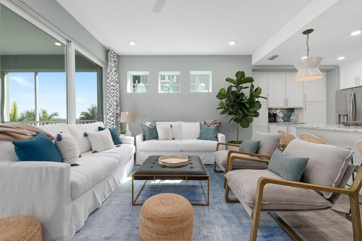 A bright living area features white sofas adorned with decorative pillows, complemented by light wooden accents. Natural light fills the space through large sliding glass doors, with a view of lush greenery in the background. A small round table sits on a soft area rug in the center.