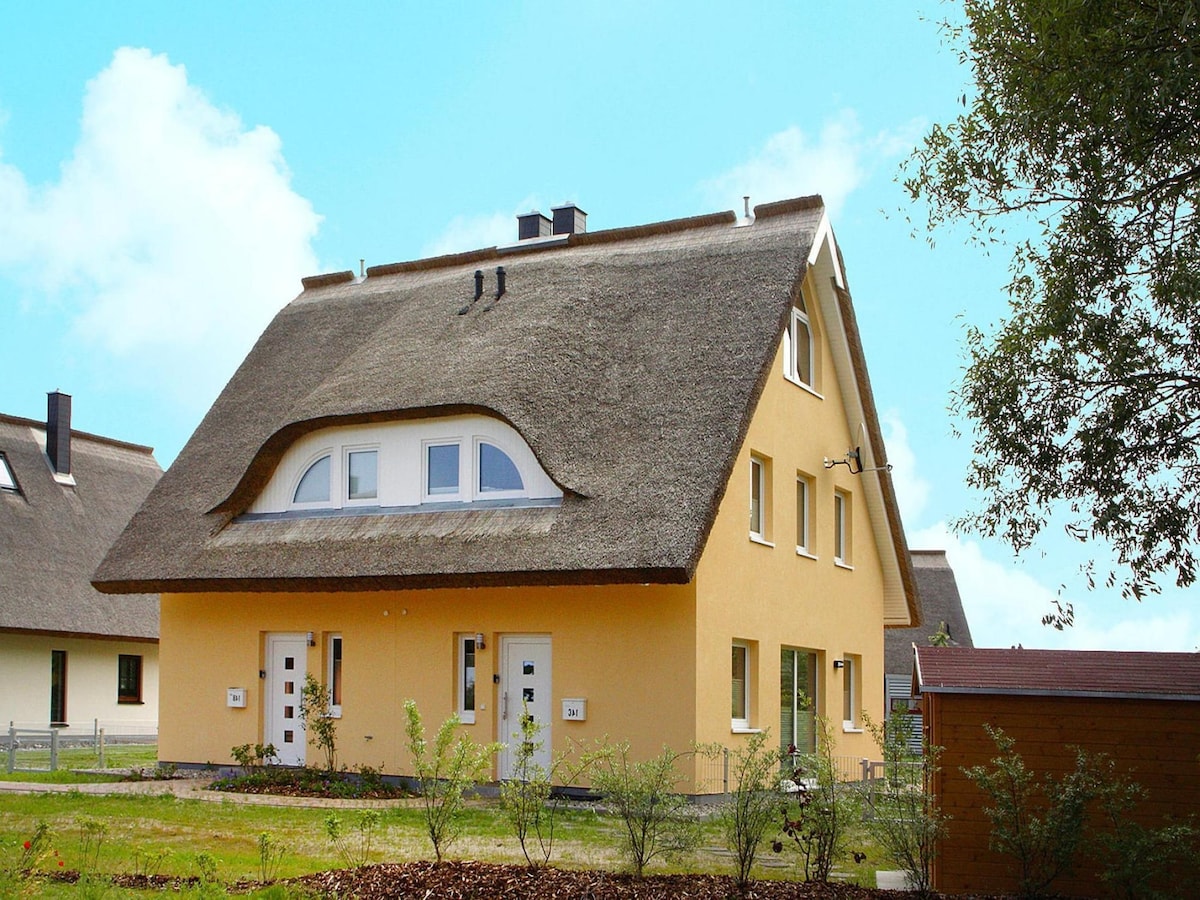 A modern two-unit house with a thatched roof is depicted. Its exterior features a warm yellow facade and curved upper window. Two front doors are visible, framed by greenery and a mulch garden bed. A nearby shed adds functionality to the property.