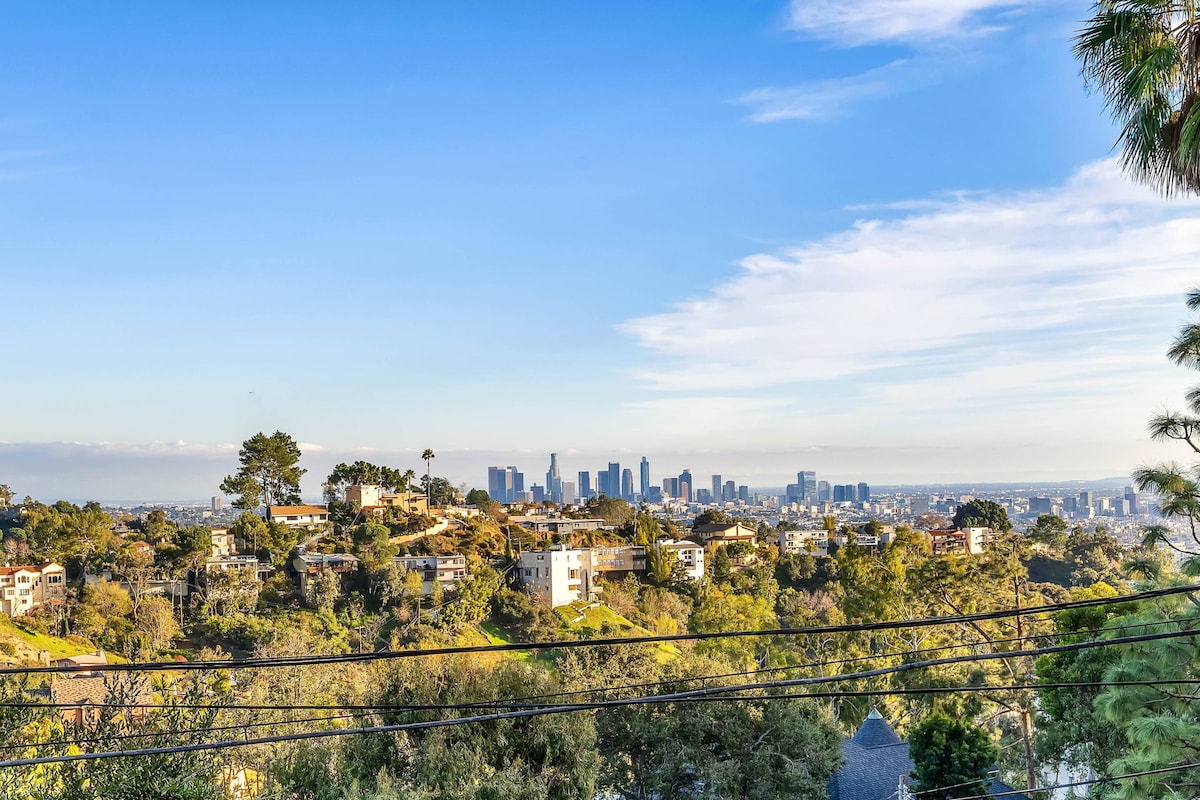 A sweeping view of downtown Los Angeles is captured against a clear sky, framed by the lush greenery of Beachwood Canyon. Houses dot the hillside, showcasing a blend of nature and urban life. Power lines run across the foreground, providing context to the striking landscape.
