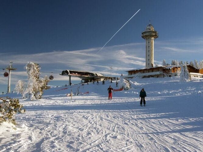 A winter scene is depicted, showcasing a snow-covered landscape with skiers on the slopes. A ski lift ascends toward a tall observation tower in the background, surrounded by a clear blue sky and scattered clouds. The ground is blanketed in fresh snow, enhancing the serene atmosphere.