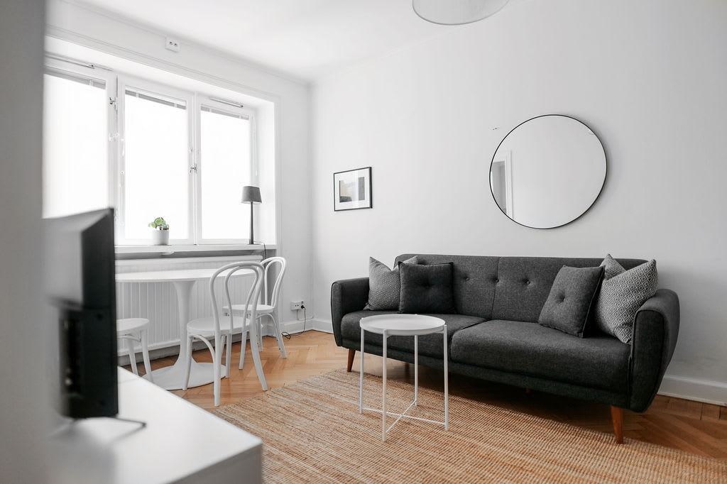 A modern living area features a dark gray sofa with decorative pillows, positioned against a bright wall. A round mirror hangs above the sofa, reflecting natural light from the windows. A small dining table with two chairs and a light interior complete the cozy space.