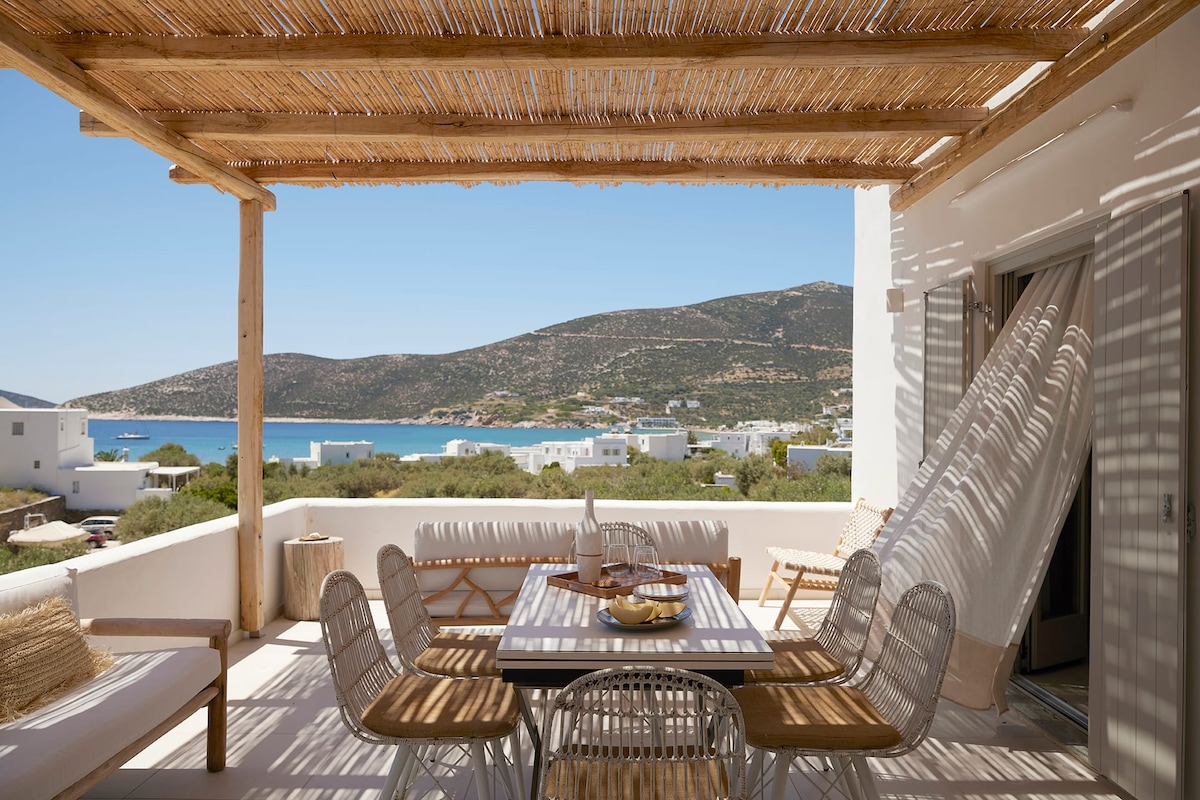 An outdoor terrace features a dining table surrounded by stylish chairs, with sunlight filtering through a bamboo pergola. Scenic views of the hills and coastline can be seen in the background, creating a serene setting for meals and relaxation.