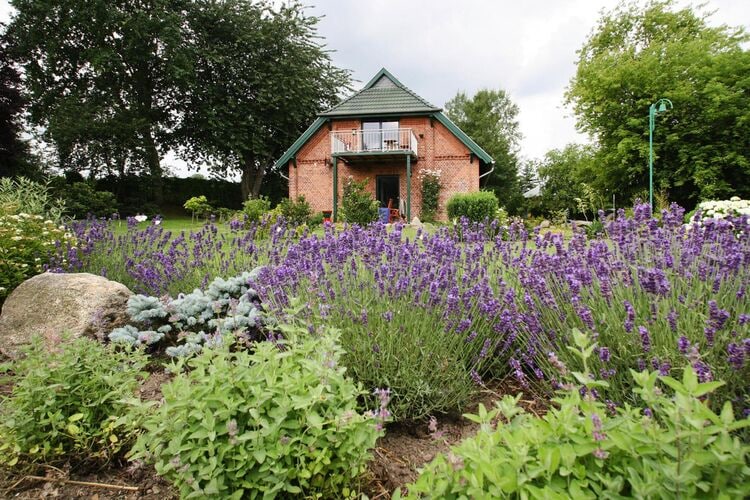 A charming two-story country house is surrounded by lush gardens, featuring vibrant lavender and green shrubs. A small rock is positioned in the foreground, adding natural texture. The balcony above invites relaxation and overlooks the beautifully maintained landscape.