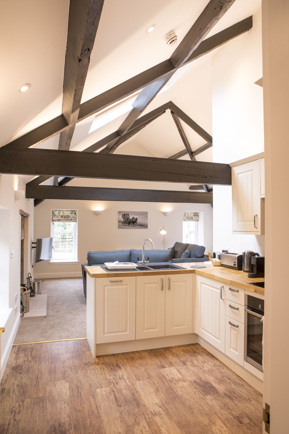 The open-plan living area showcases high ceilings accentuated by exposed oak beams. A modern kitchen features sleek cabinetry and appliances, while a cozy seating arrangement is positioned nearby, allowing for a spacious and airy atmosphere.